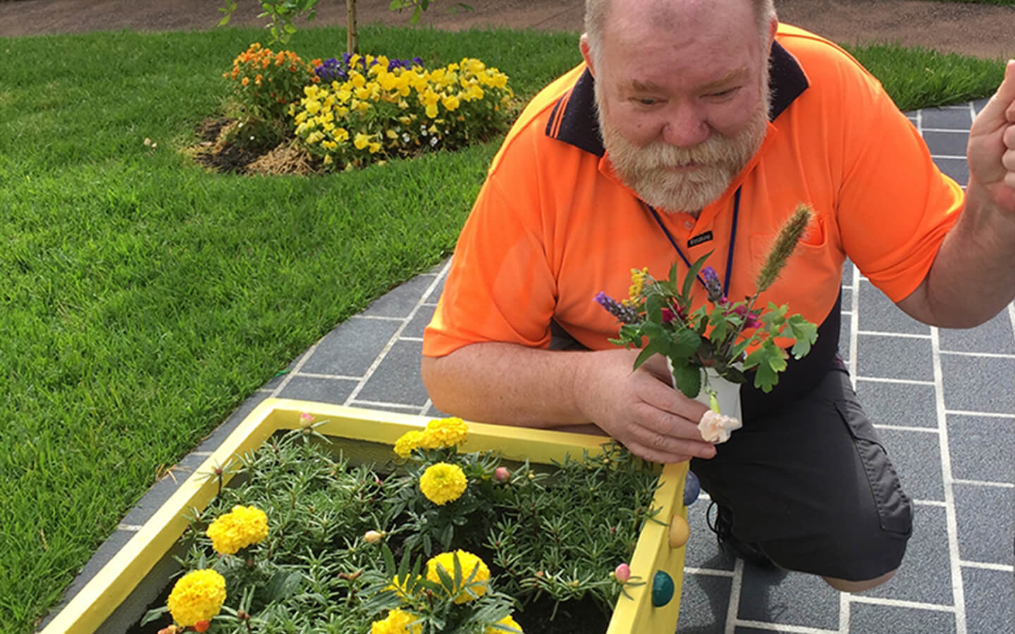 person holds a plant and crouches next to flowers in a large pot