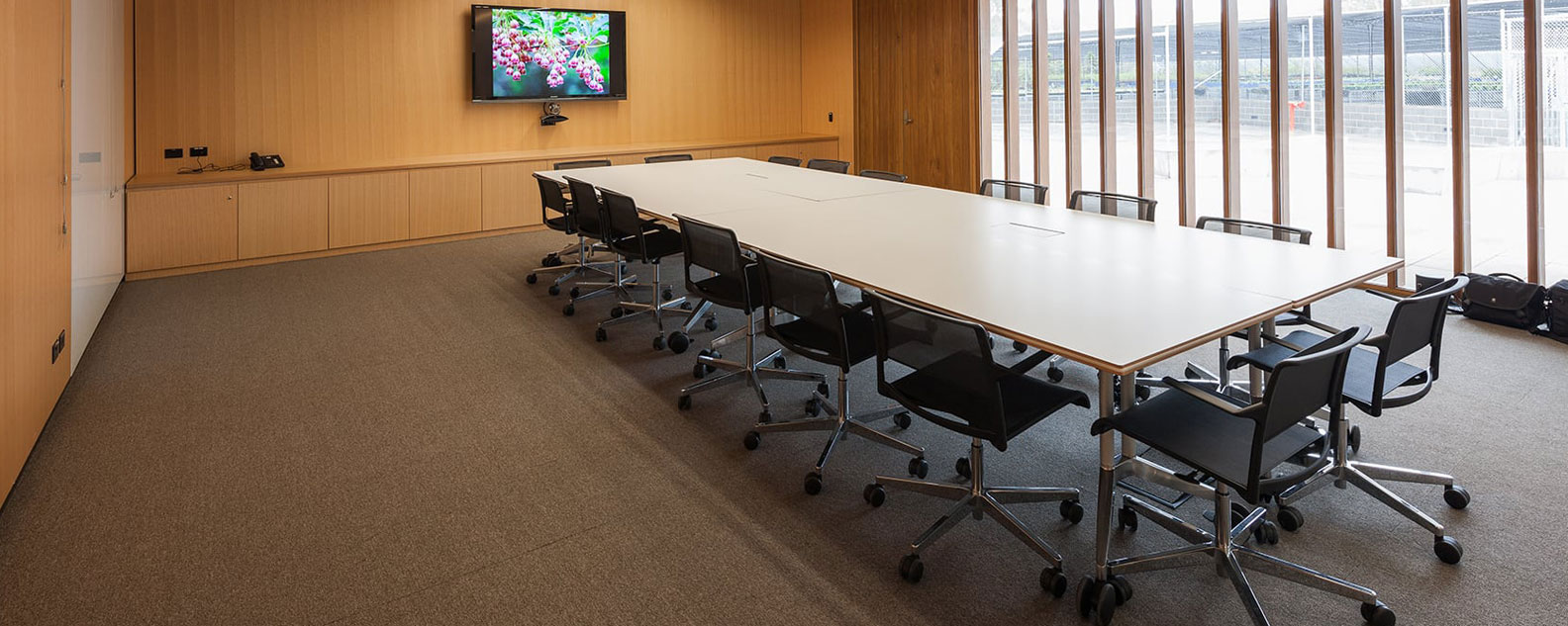 A corporate meeting room with wood panelled walls, floor to ceiling windows, a presentation screen and a large meeting table with seating.