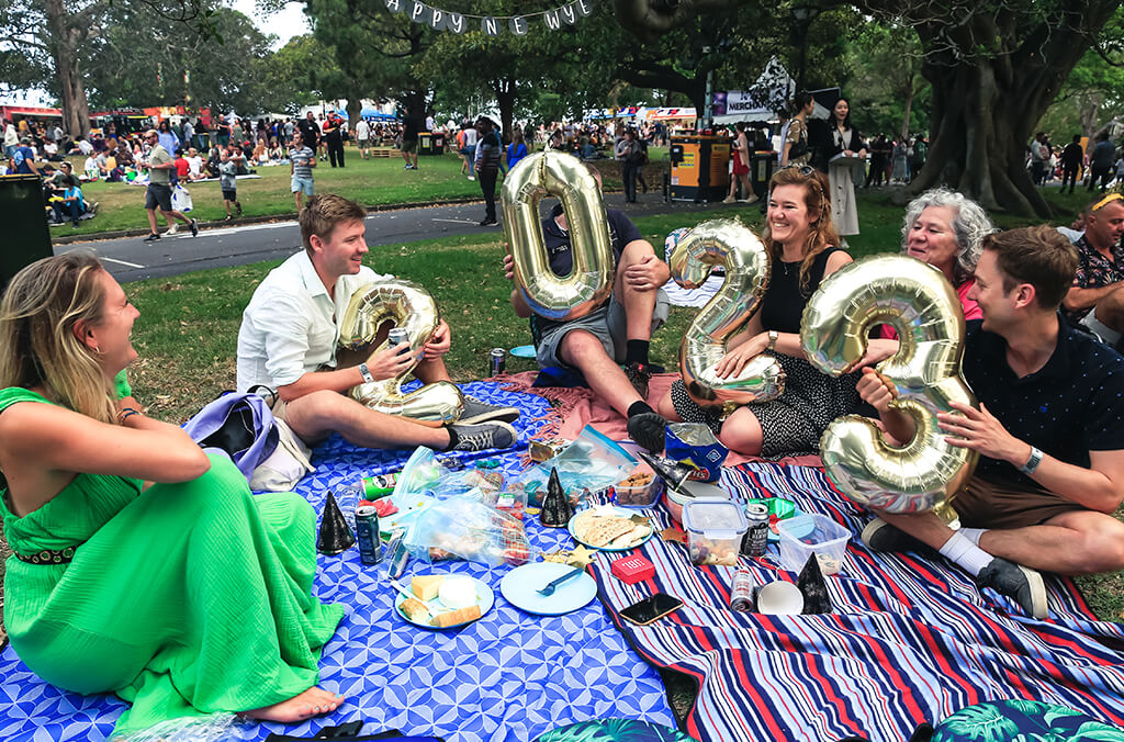 Group of picnickers on a rug in garden setting, holding 2023 balloons