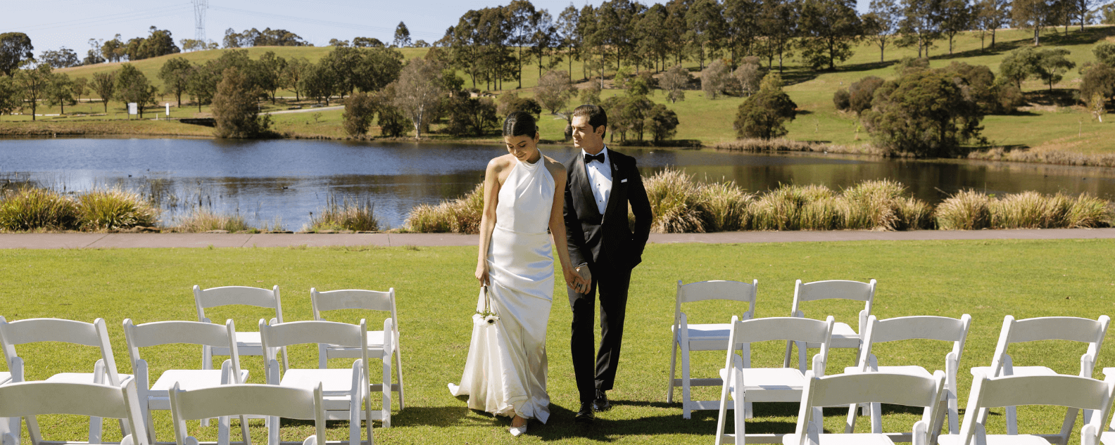 Two people walking down an aisle with a lake and garden in the background and chairs on the side