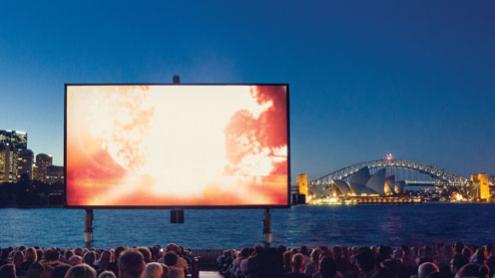 A seated crowd watch a big screen with the harbour and Sydney Harbour Bridge in the background