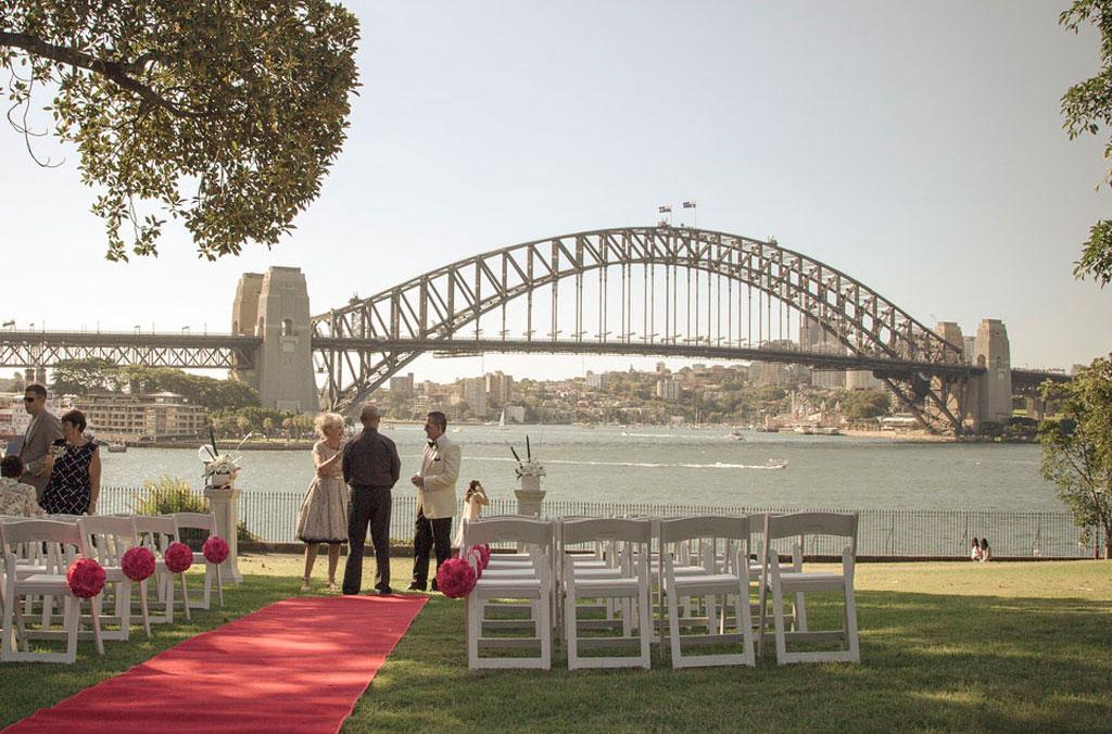 A red carpet with a couple and a celebrant with the harbour and harbour bridge in the background.