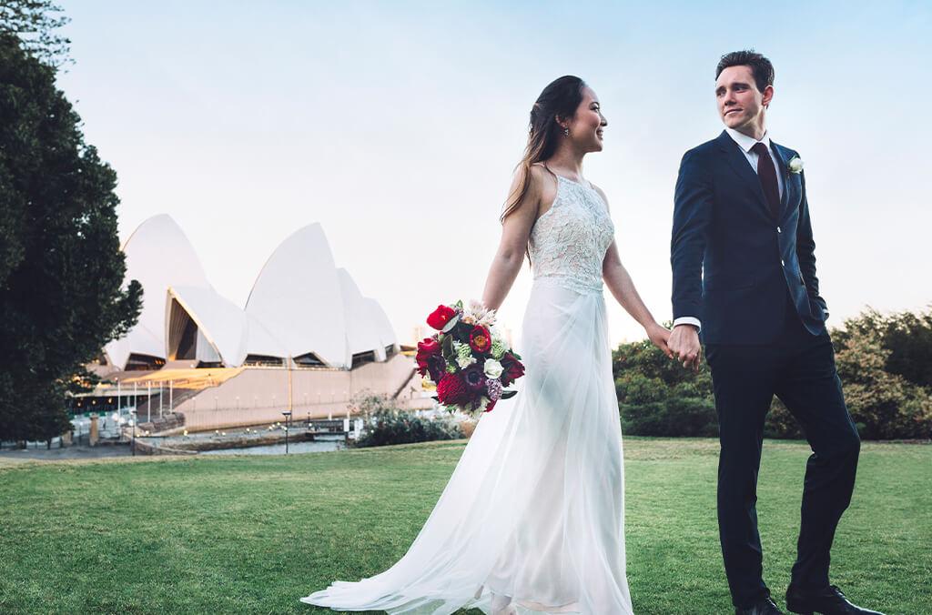 Bride and groom walk across lawn holding hands, the Sydney Opera House behind them