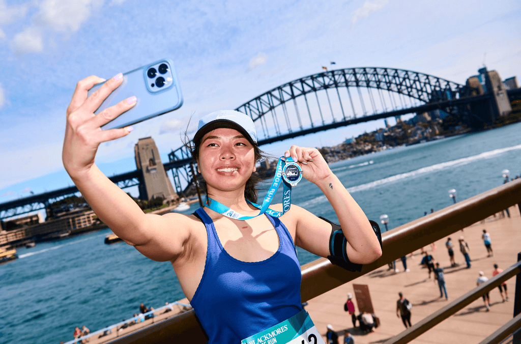 Runner takes a selfie with their medal with Sydney Harbour Bridge and shoreline in background.