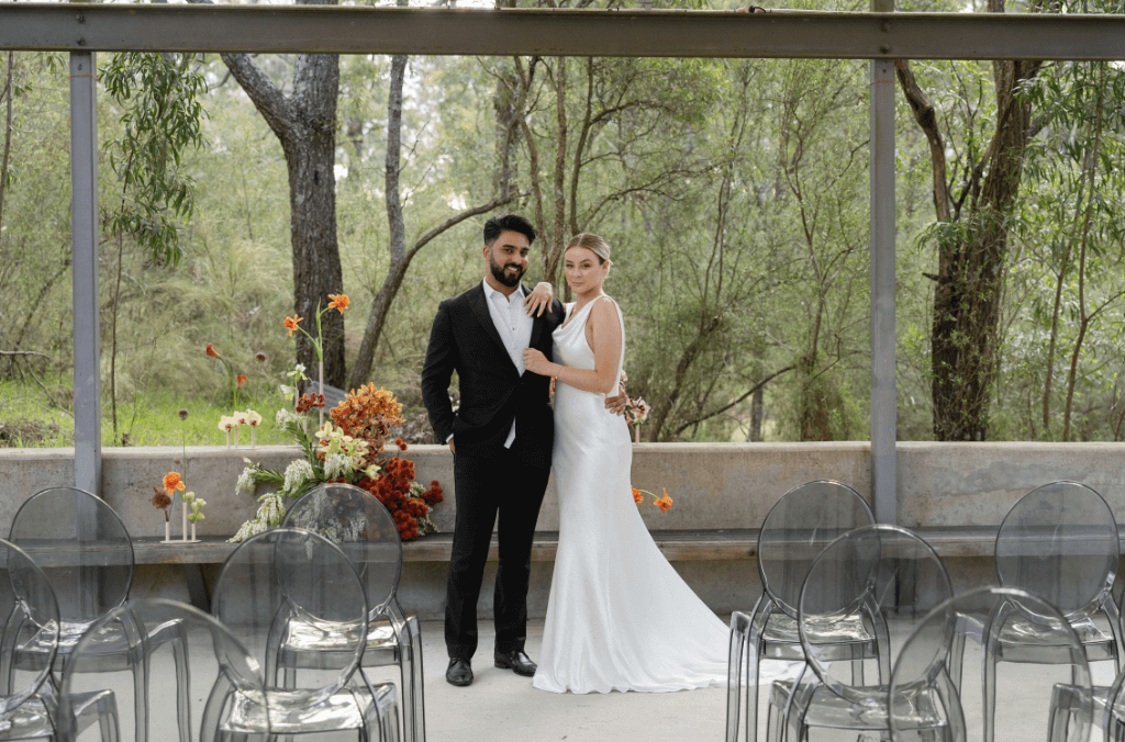 A couple standing under a shelter with clear chairs and trees in the background