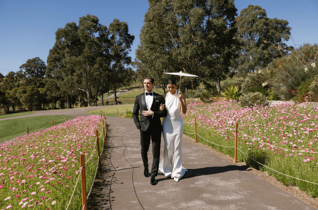 A man and women holding an umbrella walk down a garden path with pink daises on either side